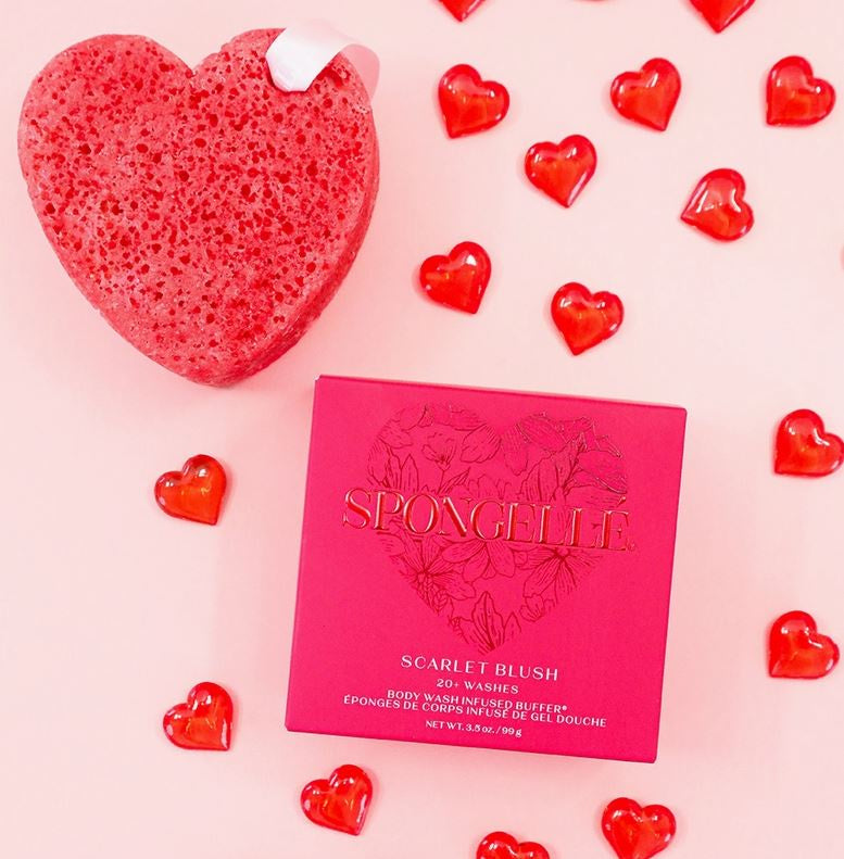 Commercial product photograph by Spongellé featuring a porous red heart-shaped body wash buffer and its gold-embossed Scarlet Blush gift box on a pink background decorated with scattered translucent red heart gems and a white ribbon loop