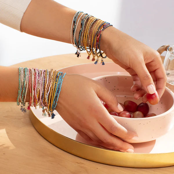 Lifestyle photograph of hands wearing stacks of colorful seed bead tassel bracelets reaching into a pink speckled bowl of red grapes on a gold-rimmed circular tray