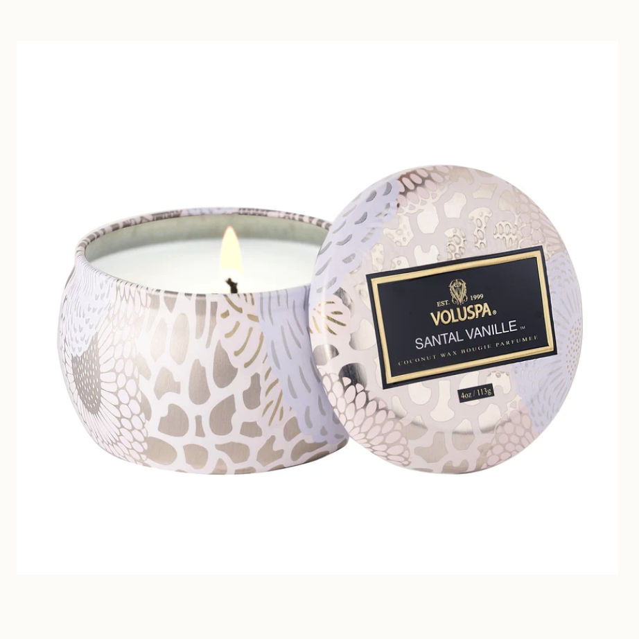 Scented coconut wax candle by Voluspa in a decorative tin with a single lit wick on a white background featuring an iconic black and gold Santal Vanille label and intricate lavender floral patterns with metallic accents