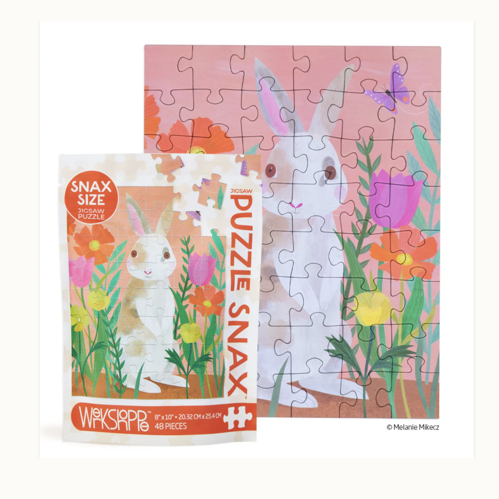 Jigsaw puzzle by artist Melanie Mikecz depicting a whimsical white and brown rabbit in a garden with vibrant pink tulips, orange poppies, and a purple butterfly against a pink background alongside its Puzzle Snax packaging