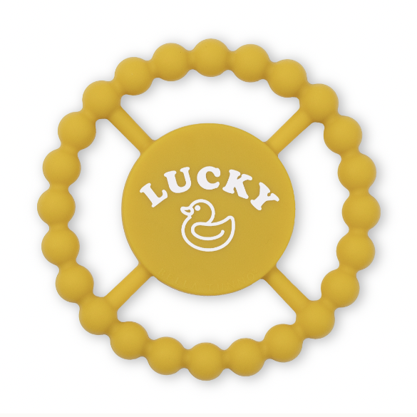 Silicone baby teether by Bella Tunno featuring a mustard yellow wheel-shaped design on a white background with a textured beaded outer ring and a central disc displaying bold white LUCKY text and a rubber duck graphic