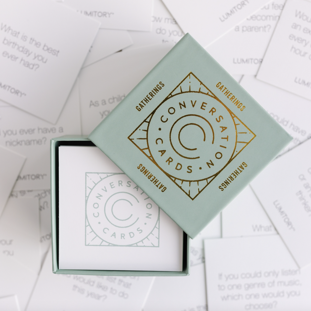 Product photograph of Lumitory Gatherings Conversation Cards featuring a sage green box with a metallic gold foil diamond logo and scattered white cards with legible icebreaker prompts like what is the best birthday you ever had