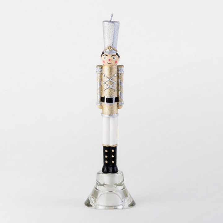 Hand-painted wax nutcracker soldier candle standing in a glass holder against a white background featuring a shimmering gold glitter uniform, a tall silver shako hat, and black boots with gold accents
