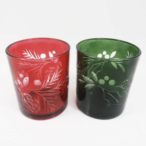 Etched glass candle holders featuring a vibrant red tumbler with frosted pinecone motifs and a dark green tumbler with holly berry etchings on a white background