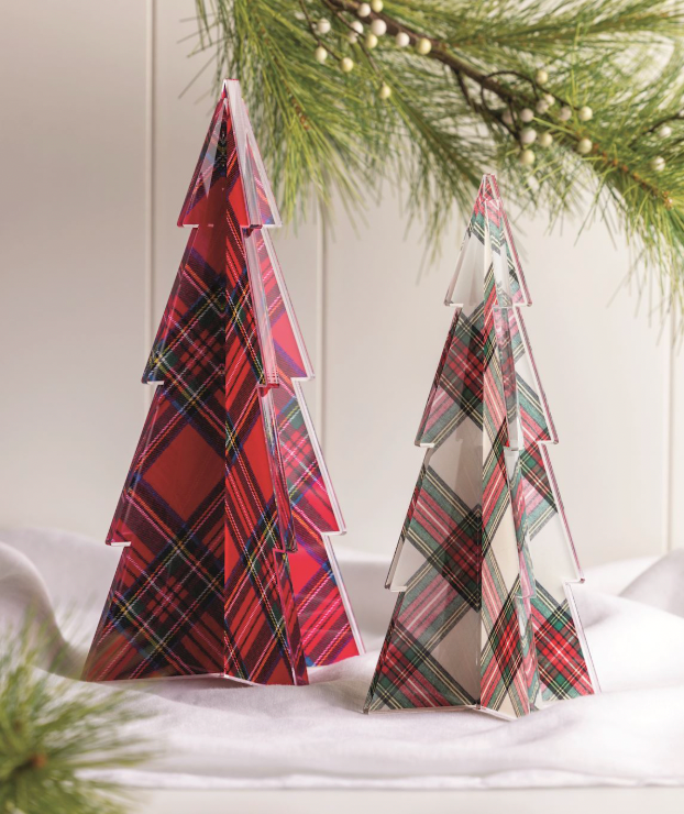 Acrylic holiday figurines of two tartan patterned Christmas tree tabletop decorations set on a white cloth beneath a natural pine branch, featuring a red Stewart plaid, a cream and green tartan, and clear translucent faceted edges