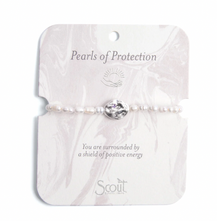 Jewelry photography by Scout Curated Wear of a Pearls of Protection bracelet featuring small white freshwater pearls and a central hammered silver charm with a tiny purple crystal on a light pink marbled display card featuring the brand logo at the bottom