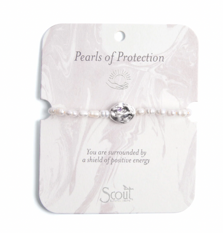 Jewelry by Scout Curated Wear featuring a delicate bracelet of small white pearls centered on a marble-patterned display card with a hammered silver charm inset with a tiny purple crystal and the text Pearls of Protection