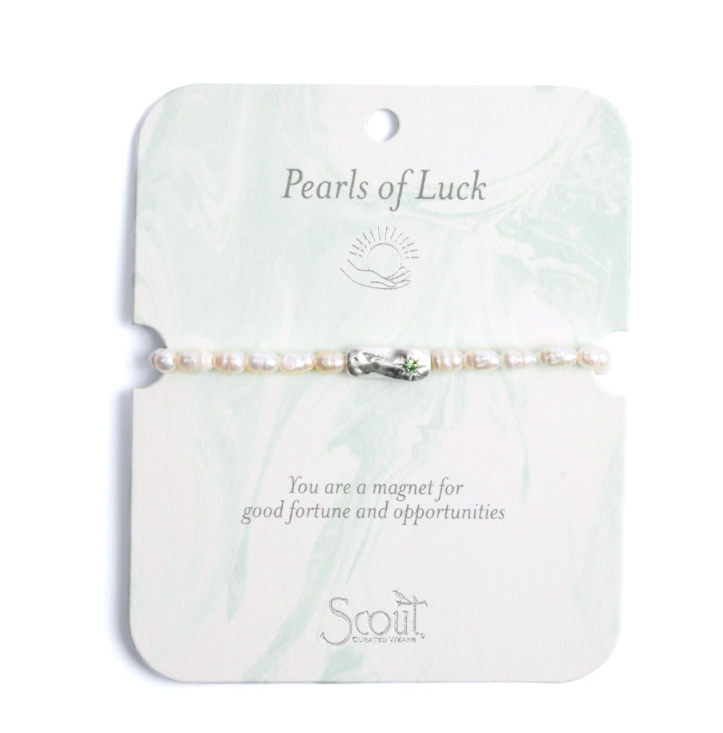 Product photography of a Scout Curated Wears bracelet centered on a marbled card featuring iridescent white pearls, a hammered silver charm with a green gemstone, and the Pearls of Luck header with the sun and hand icon