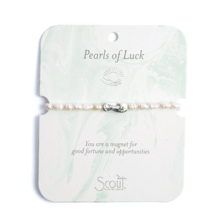 Product photography of a Scout Curated Wears bracelet centered on a marbled card featuring iridescent white pearls, a hammered silver charm with a green gemstone, and the Pearls of Luck header with the sun and hand icon