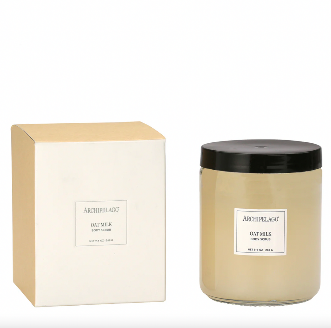 Studio photograph of Archipelago Oat Milk Body Scrub featuring its minimalist tan and white packaging box and a clear glass jar with a black screw-top lid and creamy beige contents on a white background