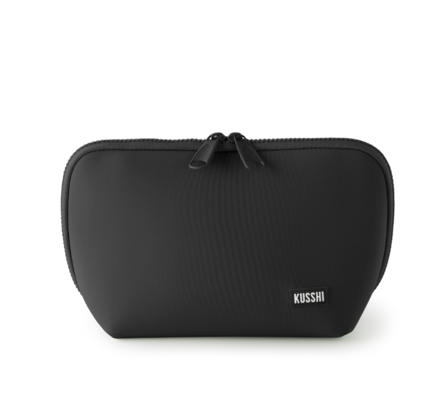 Product photograph by KUSSHI of a black travel makeup bag on a white background featuring dual black zippers, a matte fabric texture, and a small rectangular white-lettered brand patch