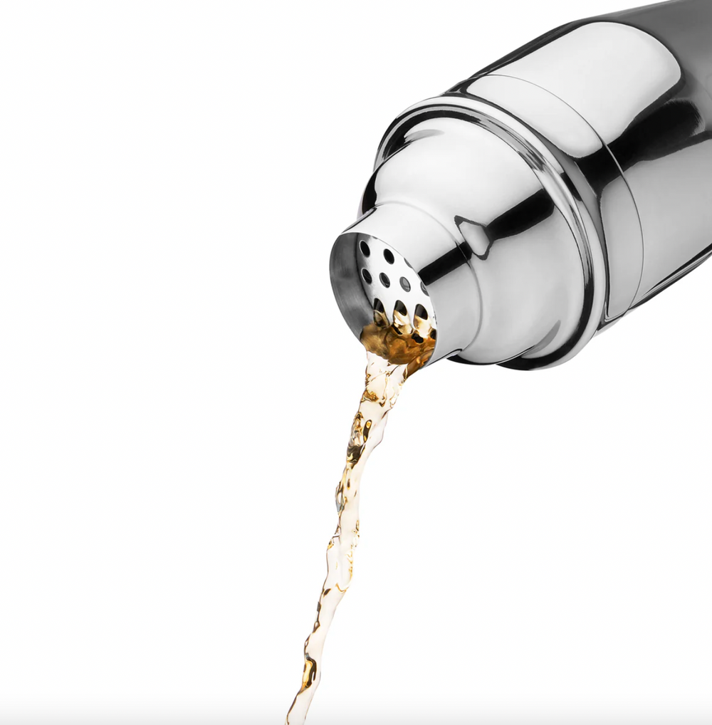 Studio photograph of a polished stainless steel cocktail shaker pouring a stream of golden amber liquid against a white background featuring a reflective chrome finish and a perforated strainer lid