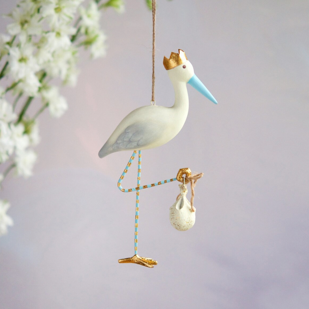 Handcrafted painted stork ornament hanging against a soft white floral backdrop, featuring a gold-crowned head, a light blue beak, and blue and gold striped legs carrying an iconic baby bundle