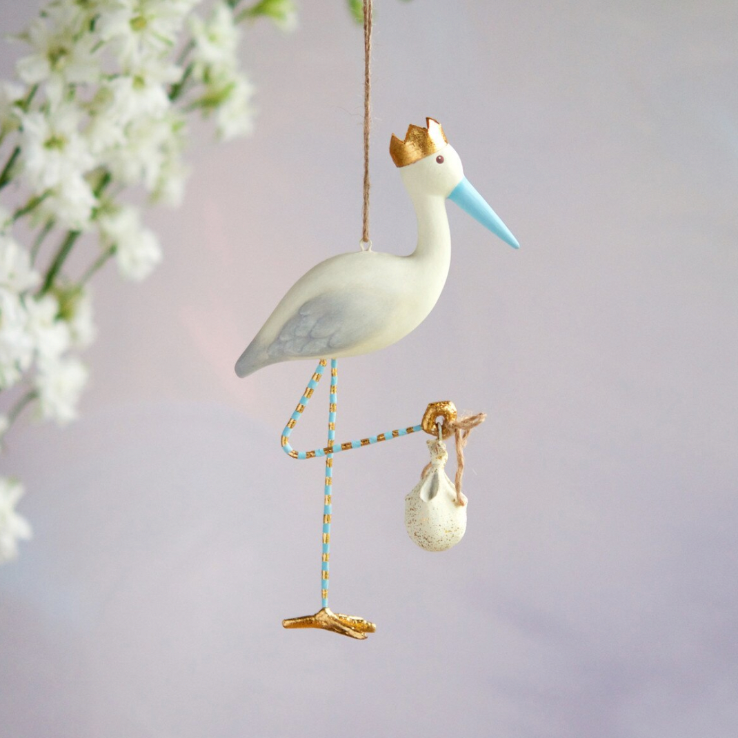 Handcrafted painted stork ornament hanging against a soft white floral backdrop, featuring a gold-crowned head, a light blue beak, and blue and gold striped legs carrying an iconic baby bundle