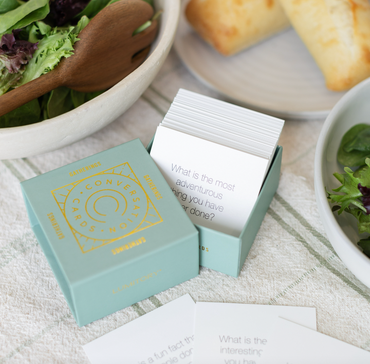Photograph of Lumitory Conversation Cards on a dining table featuring a sage green box with a gold foil circular logo next to a fresh salad with wooden servers and a card asking about adventurous experiences