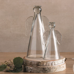 Glass sculptures of two clear angel figurines resting on stacked white birch wood discs featuring minimalist conical bodies, transparent wings, and a sprig of green leaves on a warm wooden tabletop
