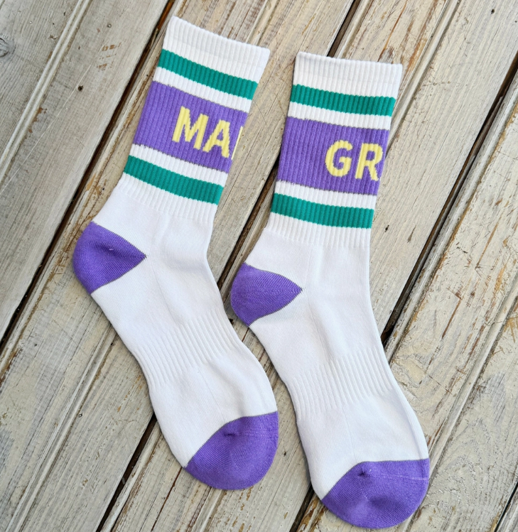 Photograph of a pair of Mardi Gras crew socks displayed on light wooden planks featuring horizontal purple and green stripes, yellow block lettering, and contrasting purple heels and toes