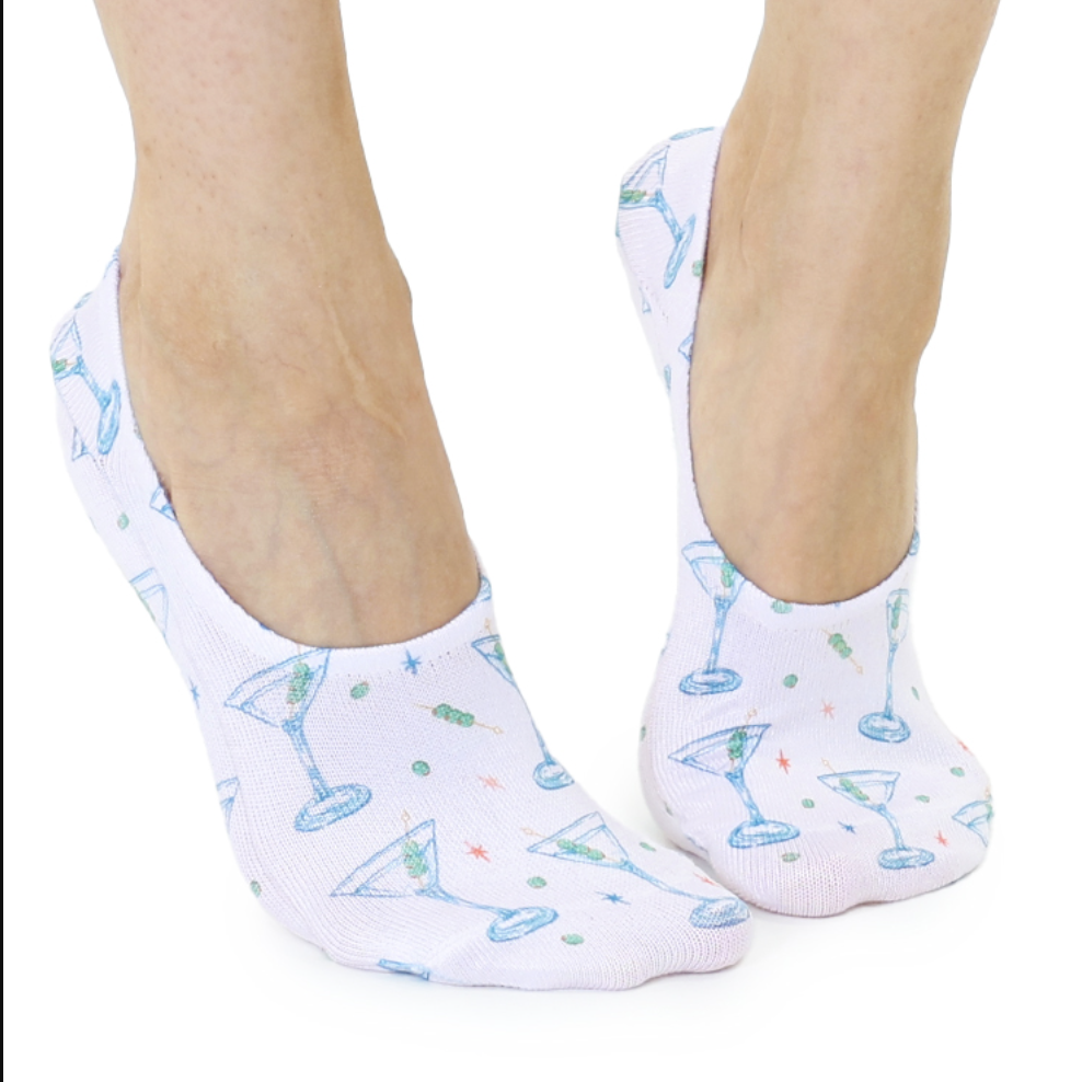 Photograph of feet wearing white no-show socks decorated with an illustrated pattern of light blue martini glasses, skewered green olives, and tiny orange starbursts against a solid white background