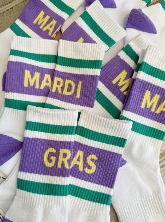 Photograph of white crew socks featuring vibrant purple and green stripes with bold yellow MARDI GRAS block lettering and a ribbed knit texture displayed on a neutral wooden background