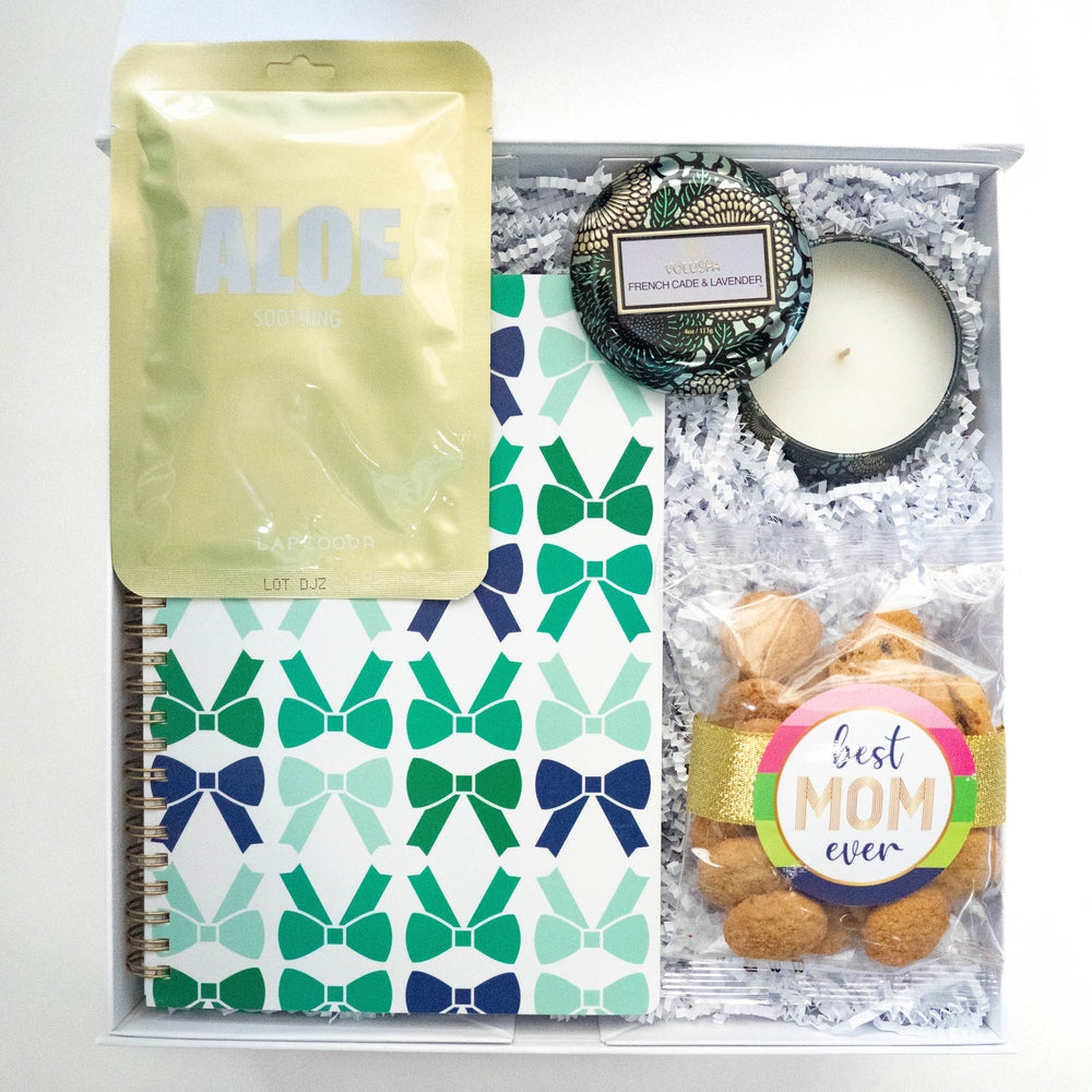 Flat-lay photograph of a Mother's Day gift box featuring a Voluspa French Cade and Lavender candle, a teal bow-patterned notebook, and a gold-packaged aloe face mask next to cookies with a Best Mom Ever label