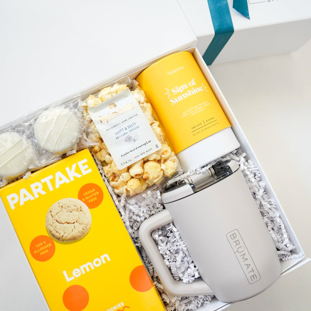 Photograph of a curated gift box featuring a light grey BrüMate insulated mug, vibrant yellow Partake Lemon cookies, and a Sips of Sunshine tea tin nestled in white crinkle paper shred