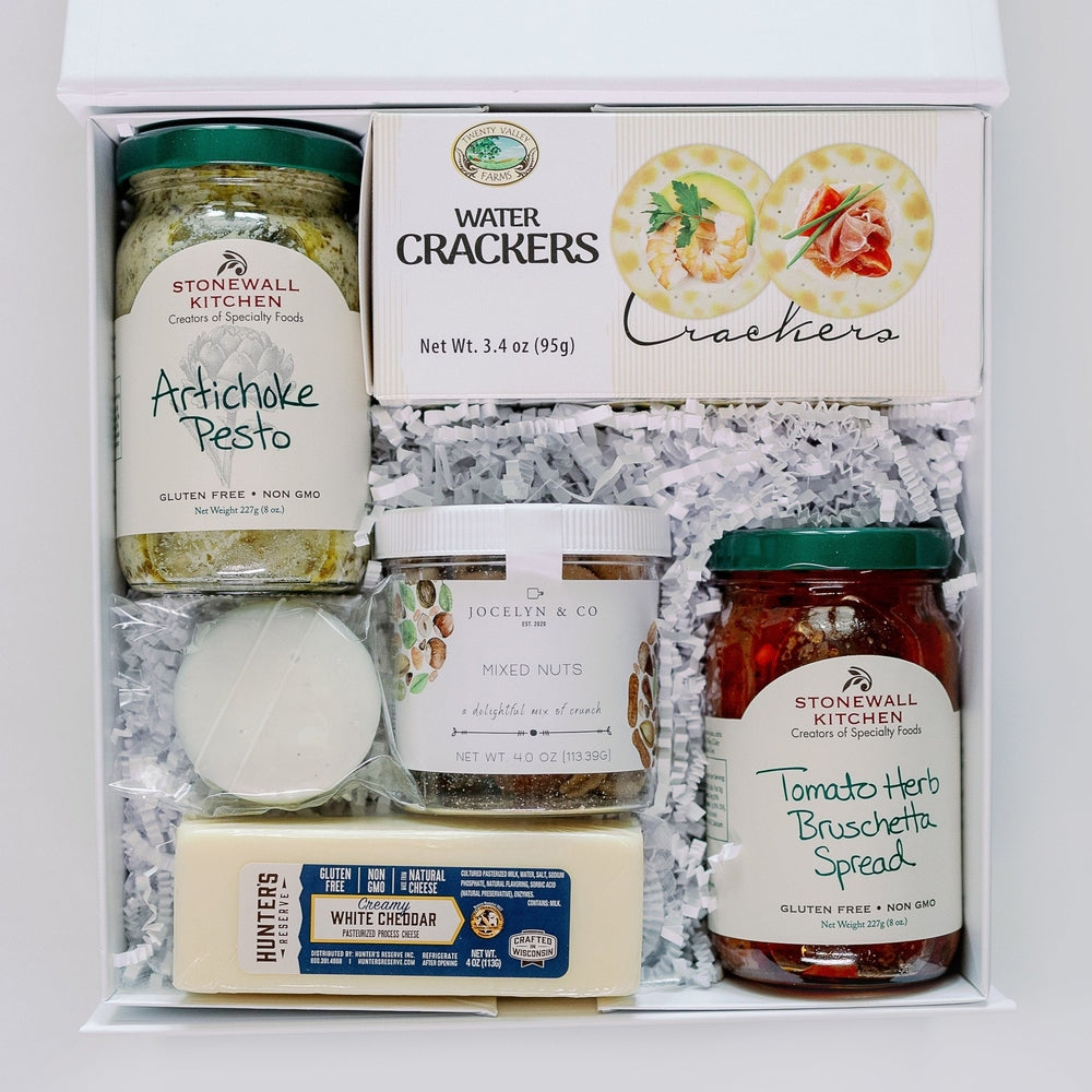 Digital photograph of a gourmet food gift box featuring green-lidded jars of Stonewall Kitchen Artichoke Pesto and Tomato Herb Bruschetta Spread nestled in white shredded paper with Twenty Valley Farms crackers and a block of creamy white cheddar