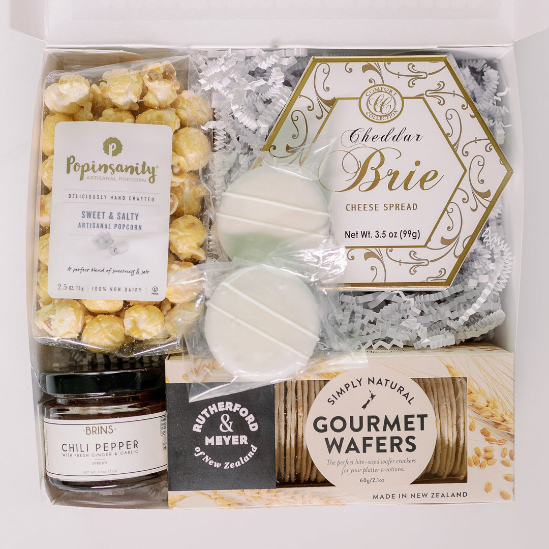 Photograph of a gourmet food gift box featuring a gold-trimmed hexagonal box of Cheddar Brie cheese spread, white-striped chocolate-covered cookies, and a clear bag of Popinsanity popcorn nestled on white shredded paper filler alongside a jar of chili pepper spread and savory gourmet wafers