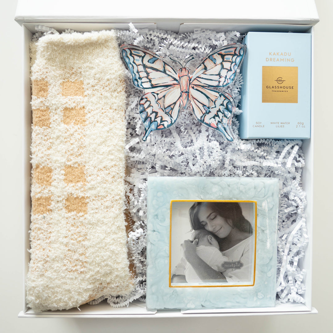 Digital photograph of a curated gift box containing a light blue Glasshouse Fragrances candle, a textured Mud Pie picture frame, plush cream-and-tan checkered socks, and a large decorative butterfly ornament on white shredded paper