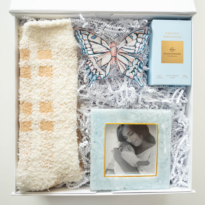 Digital photograph of a curated gift box containing a light blue Glasshouse Fragrances candle, a textured Mud Pie picture frame, plush cream-and-tan checkered socks, and a large decorative butterfly ornament on white shredded paper