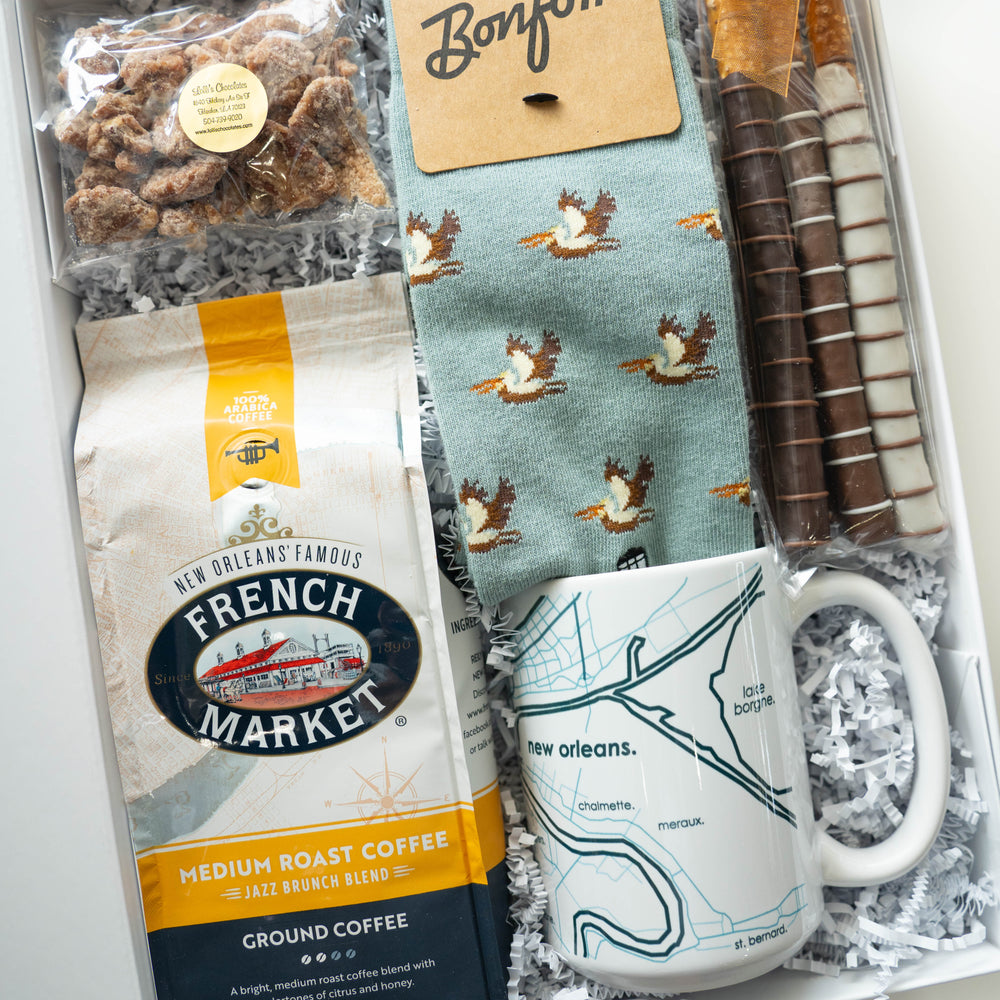 Photograph of a New Orleans themed gift box containing French Market coffee with the historic market landmark, a ceramic mug depicting a map of Lake Borgne, and light blue pelican-patterned socks alongside chocolate-covered pretzel rods