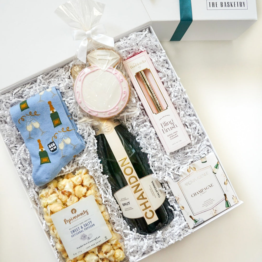 Photograph by The Basketry of a luxury celebratory gift set featuring a Chandon Brut champagne bottle, a large pink-iced diamond ring cookie, and light blue champagne-patterned socks presented in a white box with shredded paper