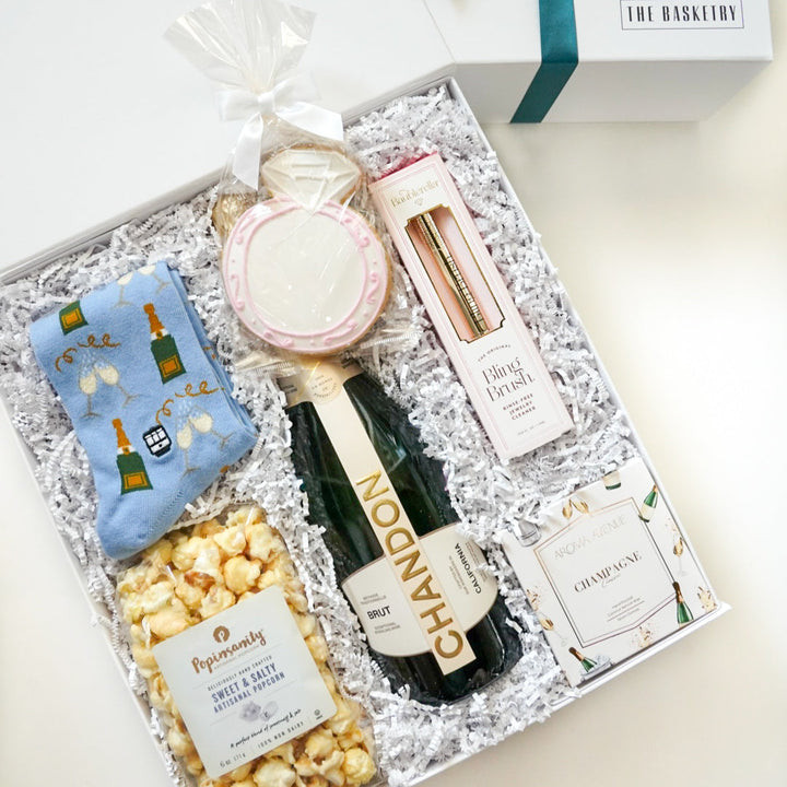 Photograph by The Basketry of a luxury celebratory gift set featuring a Chandon Brut champagne bottle, a large pink-iced diamond ring cookie, and light blue champagne-patterned socks presented in a white box with shredded paper