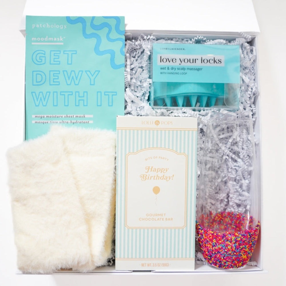 Studio photograph of a curated birthday gift box featuring a Patchology sheet mask, a Lolli & Pops chocolate bar, a teal scalp massager, and a glass flute with a vibrant rainbow sprinkle base nestled in white shredded paper