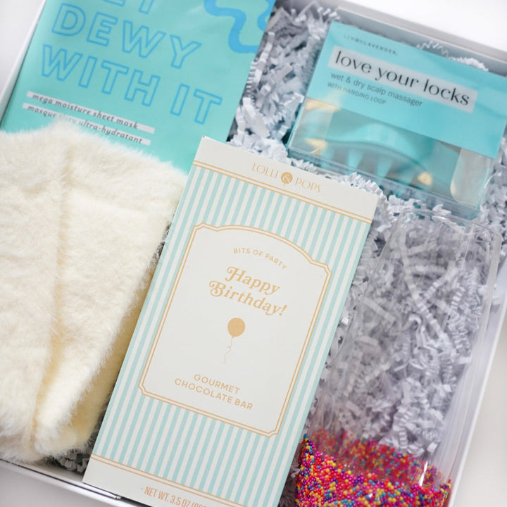 Product photograph of a curated birthday gift box featuring a Lolli &amp; Pops gourmet chocolate bar, soft white fuzzy socks, and a glass filled with vibrant rainbow sprinkles nestled in white shredded paper