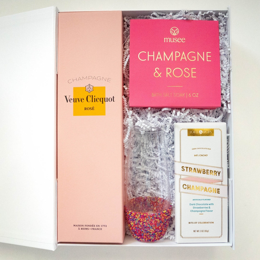 Product photograph of a luxury curated gift box featuring a pink Veuve Clicquot Rosé box from Reims France, Musee bath salts, and a stemless champagne glass with a colorful rainbow sprinkle base resting on white shredded paper filler