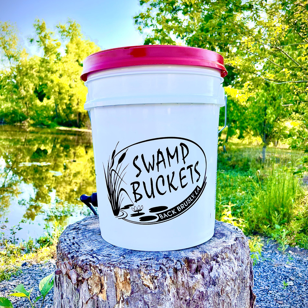 Photograph of a white Swamp Buckets pail with a vibrant red lid perched on a weathered tree stump in Back Brusly, Louisiana, featuring a black logo with cattails against a backdrop of lush green foliage and a sunlit pond