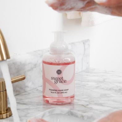 Photograph of Sweet Grace foaming hand soap on a white marble bathroom counter featuring a brushed gold faucet with running water and bubbly pink liquid inside the clear bottle with white soap suds on hands above