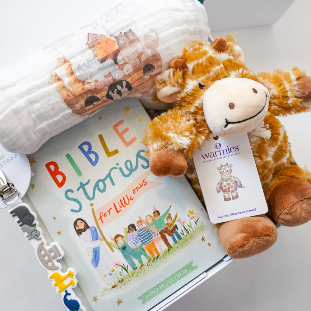 Photograph of a baby gift set by Paper Peony Press and Warmies featuring the Bible Stories for Little Ones book with rainbow lettering, a plush giraffe, a Noah's Ark printed muslin swaddle, and a pacifier clip with colorful animal charms