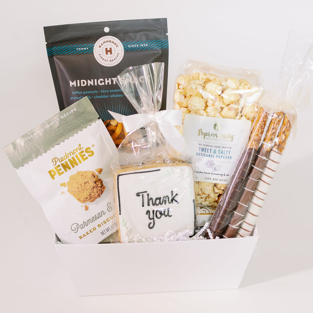 Studio photograph of a white gourmet gift basket featuring Piedmont Pennies, Hammond's Midnight Snack, and Popinsanity brands showcasing a hand-iced Thank You cookie with a white bow and multi-toned striped chocolate pretzel rods