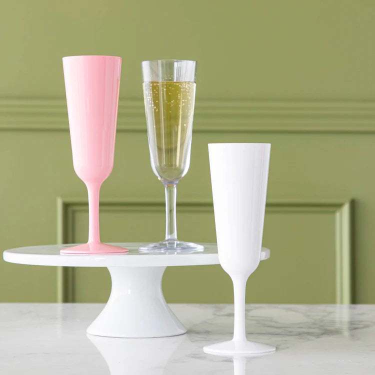 Digital photograph of three champagne flutes featuring opaque pink, solid white, and clear styles filled with bubbling golden liquid arranged on a white pedestal stand against a sage green paneled wall