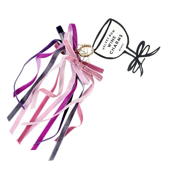 Studio product photograph of Joanna Buchanan velvet bow wine charms featuring six long ribbons in shades of pink, purple, and mauve dangling from gold-tone rings on a wine-glass-shaped packaging card