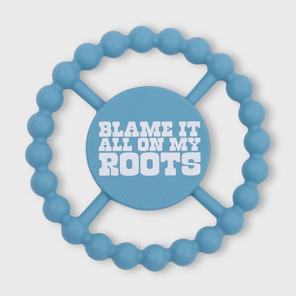 Silicone baby teether by Bella Tunno featuring a sky blue circular design with a beaded outer edge, four connecting spokes, and a central medallion displaying the words BLAME IT ALL ON MY ROOTS in white block font against a neutral background