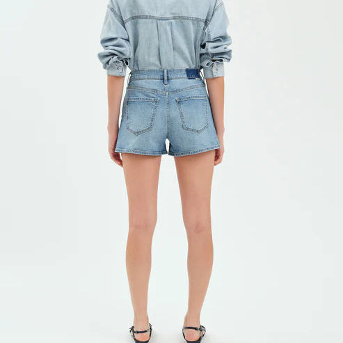 Fashion photograph of a model from behind wearing light wash denim shorts and a matching button-down shirt against a plain white studio background featuring classic back pockets and minimalist black strappy sandals