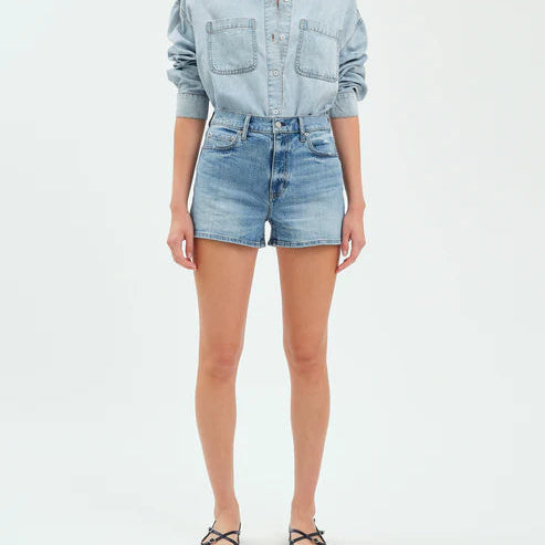 Studio fashion photograph of a model wearing a light-wash denim shirt with double chest pockets and high-waisted blue shorts against a plain white studio backdrop featuring black strappy sandals