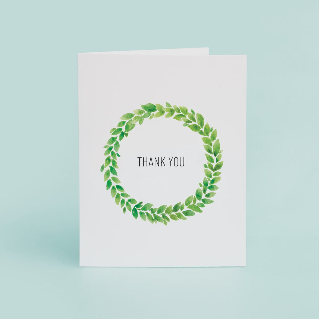 Watercolor painted greeting card featuring a circular wreath of vibrant green leaves and minimalist black THANK YOU text on a white card against a light blue background