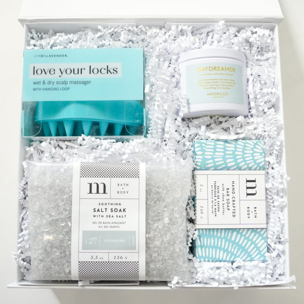 Commercial product photography of a curated self-care gift box featuring a teal Lemon Lavender scalp massager, a white Moodcast candle, and a bag of cashmere sea salt soak nestled in white crinkle paper