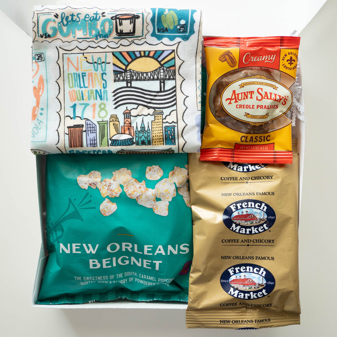 Photograph of a New Orleans-themed gift box featuring an illustrated tea towel depicting the St. Louis Cathedral, the Crescent City Connection bridge, and the Superdome alongside a teal beignet popcorn bag, a gold French Market coffee packet, and a classic Aunt Sally’s praline