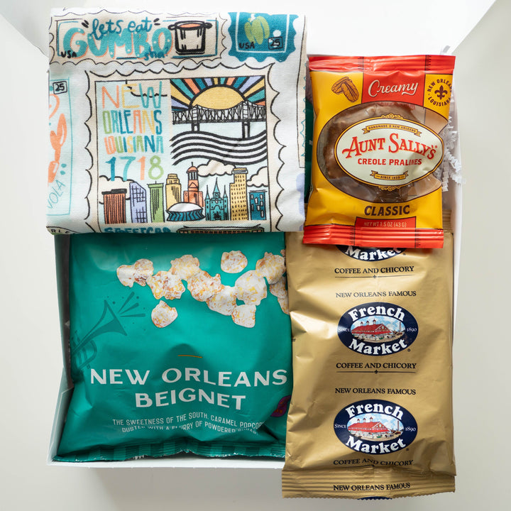 Photograph of a New Orleans-themed gift box featuring an illustrated tea towel depicting the St. Louis Cathedral, the Crescent City Connection bridge, and the Superdome alongside a teal beignet popcorn bag, a gold French Market coffee packet, and a classic Aunt Sally’s praline