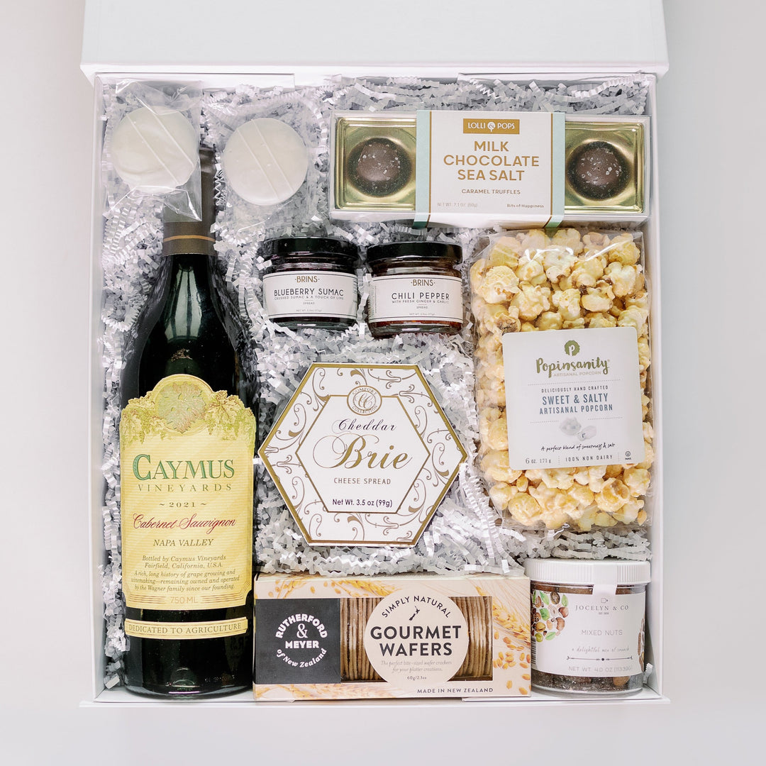 Commercial photograph of a luxury gourmet gift box containing a bottle of Caymus Vineyards Cabernet Sauvignon, Popinsanity artisanal popcorn, and a hexagonal box of Cheddar Brie cheese spread nestled in white crinkle paper with colorful jars of blueberry sumac and chili pepper jam