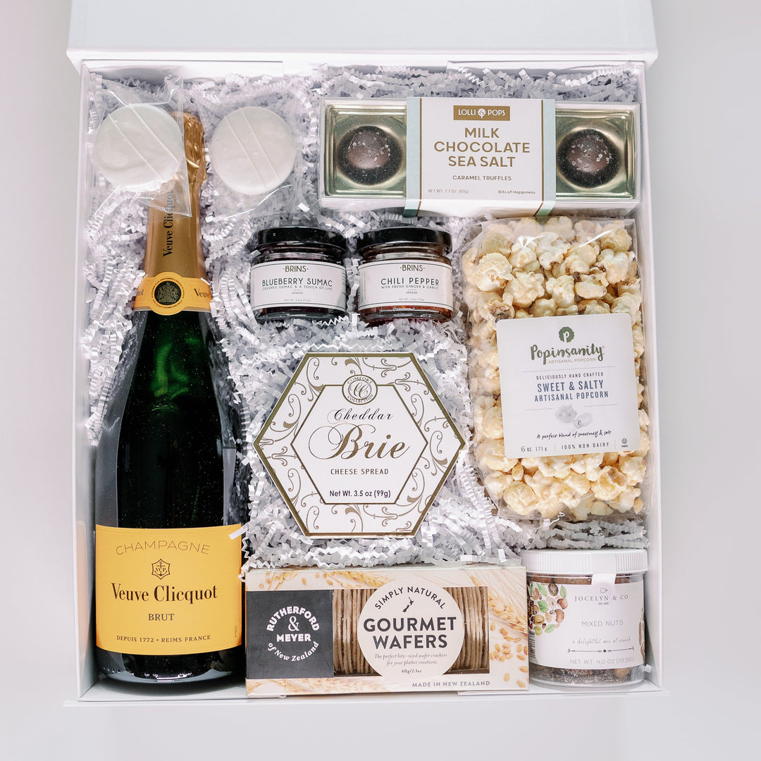 Studio photograph of a luxury gourmet gift box featuring a bottle of Veuve Clicquot Brut Champagne alongside artisanal popcorn and a gold-trimmed hexagonal cheddar brie box nestled in white shredded paper
